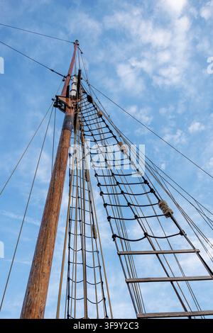Una vista ad angolo basso che guarda verso l'alto di un alto albero in legno su una nave classica. Cavi di sartiame dettagliati, funi e una scala di corda tradizionale si estendono in avanti Foto Stock