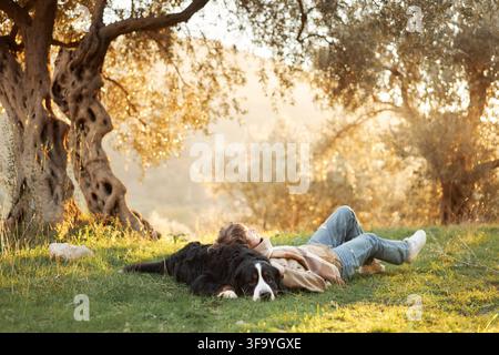 Donna si rilassa sull'erba verde accanto a un cane di montagna del Bernese durante una giornata autunnale soleggiata. Un ulivo antico e una luce dorata della campagna creano una calma Foto Stock