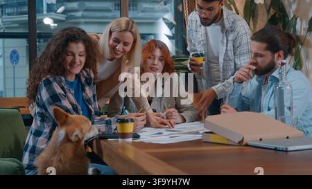 Cinque persone d'affari, squadra di lavoro informale che lavora in uno startup con caffè, pizza e cane domestico in ufficio, parlando, sorridendo e discutendo di carta, multietnico Foto Stock