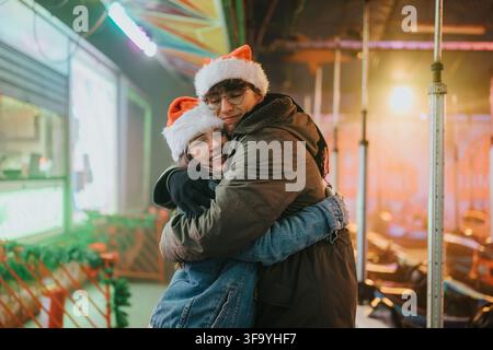 Giovane coppia che si abbraccia a un carnevale di vacanza indossando cappelli di Babbo Natale e caldi cappotti invernali Foto Stock