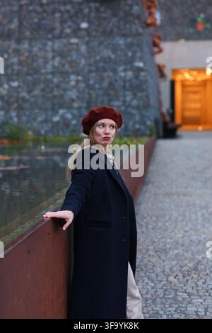 Giovane donna elegante con berretto rosso e cappotto scuro che posa all'aperto vicino a una piscina riflettente in un'architettura urbana moderna. Foto Stock