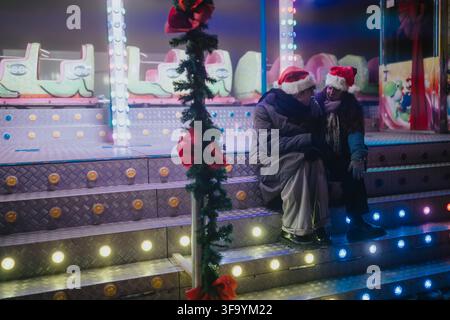 Coppia con cappelli da Babbo Natale seduta sui gradini illuminati di un'attrazione festiva di notte Foto Stock