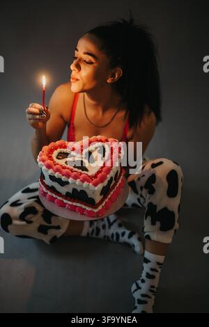 Giovane donna che tiene una torta di compleanno a forma di cuore e accende una candela mentre è seduta in pigiama Foto Stock