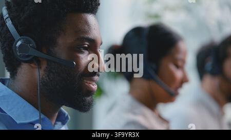 Primo piano di un receptionist afroamericano sorridente che parla in un microfono auricolare, uomo d'affari etnico spensierato, uomo d'affari al telefono in un call center Foto Stock