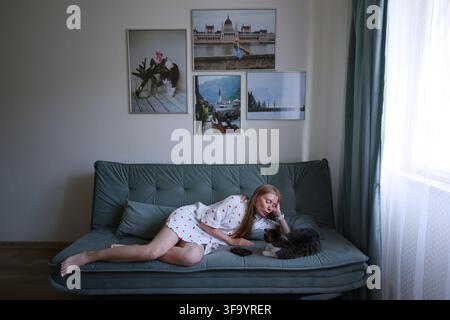 Donna sdraiata sul divano che utilizza lo smartphone mentre passa il tempo con il suo gatto in un soggiorno accogliente. Foto Stock