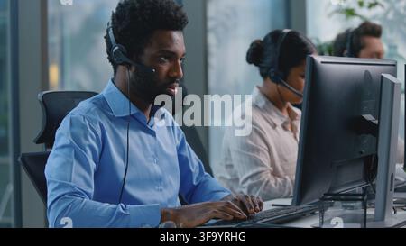 Ottimista e sorridente manager afroamericano nel call center consulente etnico addetto alla reception che parla in cuffia microfono digitando tastiera per computer Foto Stock