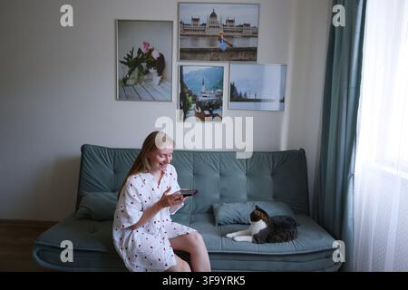 Giovane donna che controlla i messaggi sul telefono cellulare a casa mentre passa del tempo tranquillo con il suo gatto domestico. Foto Stock