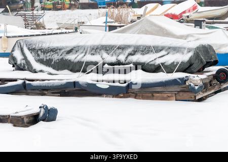 Una barca coperta da un telone è ormeggiata su un molo innevato tra altre piccole barche in una marina invernale. Foto Stock