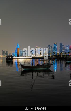 Doha, Qatar - 11 novembre 2025: Una vista verticale notturna di dhow tradizionali che galleggiano su un mare calmo vicino alla scultura 7 al MIA Park, con Foto Stock