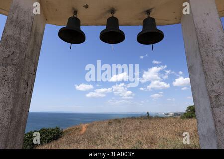 Tre campane di metallo appese all'interno di un campanile di cemento che incorniciano una vista panoramica sul vasto mare blu e sulla lontana costa sotto un cielo luminoso con dispersione Foto Stock