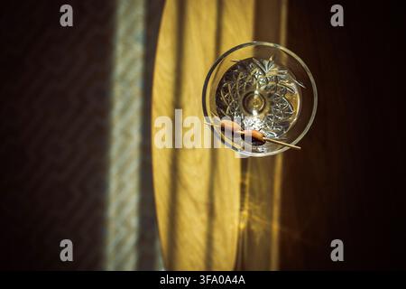 Ciotola di vetro trasparente con design intricato contiene due mandorle su un tavolo di legno, con la luce del sole che crea ombre e riflessi sulla superficie Foto Stock
