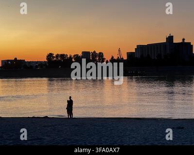 Coppia romantica che osserva il tramonto alla spiaggia di Oficirac sul Danubio, con riflessi dorati e uno skyline industriale a Novi Sad, Serbia. Foto Stock