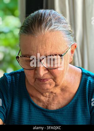 Donna anziana con occhiali guarda verso il basso, concentrata sul compito. I suoi capelli sono grigi, raccolti ordinatamente. Indossa una camicia turchese strutturata. Lo sfondo mostra una morbida na Foto Stock