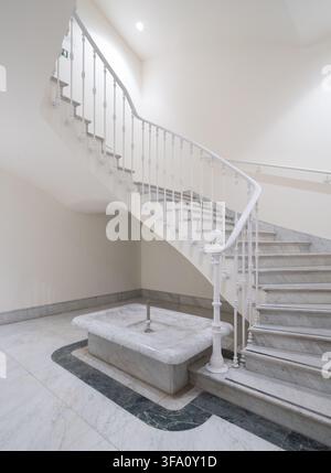 Interno di ufficio neoclassico elegante con una scala in marmo curva, ringhiera in metallo bianco ornata, pavimento in pietra e bacino della fontana centrale in un bri Foto Stock