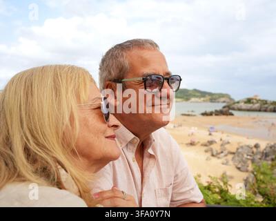 Coppia di anziani sorridenti con occhiali da sole si rilassa insieme sopra una spiaggia sabbiosa, godendosi una tranquilla vacanza costiera con vista sul mare, luce calda del giorno e l Foto Stock