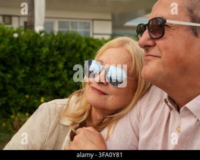 Coppia di anziani sorridenti con occhiali da sole che si rilassano insieme all'aperto, con la donna che si appoggia affettuosamente al partner in un tranquillo giardino residenziale Foto Stock