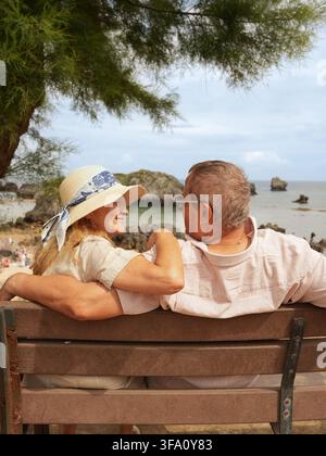 Coppia di anziani si rilassa insieme su una panchina di legno con vista su una spiaggia sabbiosa e un mare calmo, condividendo un momento affettuoso durante una tranquilla vacanza costiera Foto Stock