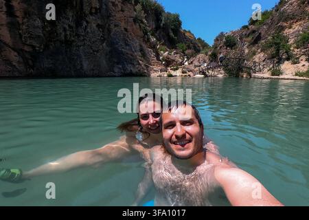 Coppia sorridente che si scatta un selfie nuotando in una piscina naturale turchese circondata da scogliere rocciose e vegetazione mediterranea a Bolbaite, Valencia Foto Stock