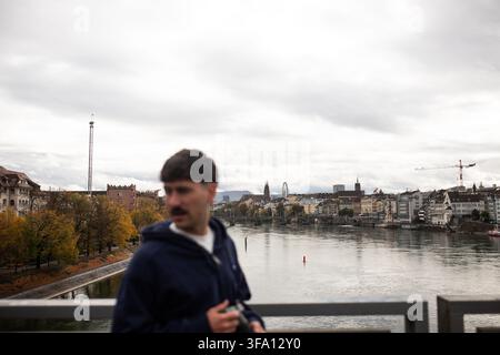 Un viaggiatore si trova su un ponte che si affaccia sul Reno e sull'antico paesaggio urbano di Basilea in Svizzera, con alberi autunnali, edifici sul lungofiume, nuvoloso Foto Stock