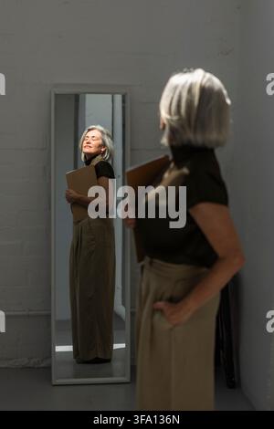 Donna matura in abbigliamento da ufficio elegante tiene una cartella mentre sorride al suo riflesso in uno specchio a figura intera, trasmettendo fiducia, accettazione di sé, a Foto Stock