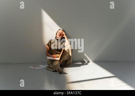 Donna d'affari matura seduta sul pavimento con una cartella aperta e fogli accanto, una mano appoggiata sulla fronte in uno spazio di lavoro minimalista illuminato dal sole. Foto Stock