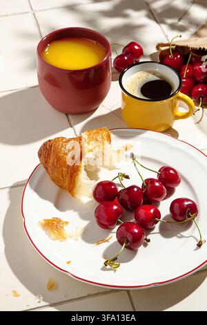 Natura morta minimalista di caffè nero, succo d'arancia, croissant friabile e ciliegie mature disposti su un piatto bianco e un tavolo piastrellato nella calda luce naturale Foto Stock