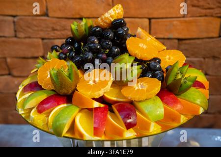 Vassoio di frutta fresca splendidamente disposto con mele, arance, kiwi e uva nera su uno sfondo di muro di mattoni. Catering gourmet salutare Foto Stock