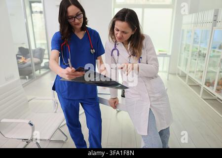 Due dottoresse esaminano insieme una radiografia toracica in un luminoso ufficio ospedaliero moderno, indossando camici da infermiera e camici da laboratorio mentre discutono della diagnosi accanto Foto Stock