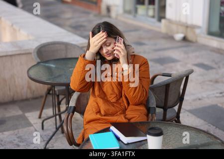 Giovane donna stressata in cappotto arancione seduta in un caffè all'aperto, tiene uno smartphone all'orecchio con una mano sulla fronte accanto a un taccuino, un laptop e una tazza di caffè Foto Stock