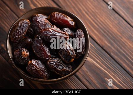 Datteri marrone scuro rugosi ammucchiati in una ciotola rustica, presentati su uno sfondo di tavola di legno texture. Questa immagine mette in mostra la dolcezza naturale e la salute Foto Stock