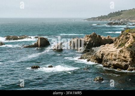 Grandi formazioni rocciose frastagliate si ergono dall'oceano lungo una costa nebbiosa, con le onde che si infrangono sulla riva Foto Stock