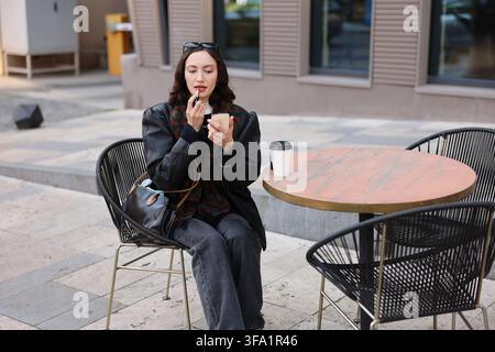 Giovane donna che applica il rossetto seduta all'aperto a un tavolo di un caffè urbano con una borsa a mano e una tazza di caffè accanto a lei nella luce soffusa del giorno, stile casual elegante Foto Stock