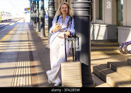 Donna matura in attesa del treno sul binario, passa il tempo con lo smartphone accanto alla valigia con le ruote. Concetto di viaggio d'affari in solitaria e stile di vita digitale Foto Stock