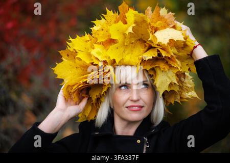 Un ritratto in primo piano di una donna bionda pensierosa in giacca nera. Tiene sulla testa una grande corona di foglie autunnali giallo brillante contro un Foto Stock