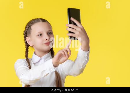 Simpatica preadolescente con trecce in camicia bianca tiene uno smartphone, facendo un selfie o una videochiamata. Bambino moderno che utilizza la tecnologia. Scatto in studio su vibrante Foto Stock