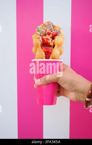 un bubble waffle servito in una tazza di carta rosa brillante su uno sfondo a righe rosa e bianche Foto Stock