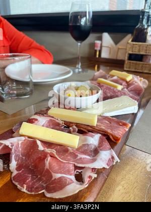Una selezione di carni affumicate e vari formaggi serviti su una tavola di legno, con un bicchiere di vino rosso sullo sfondo del ristorante. Thailandia Foto Stock