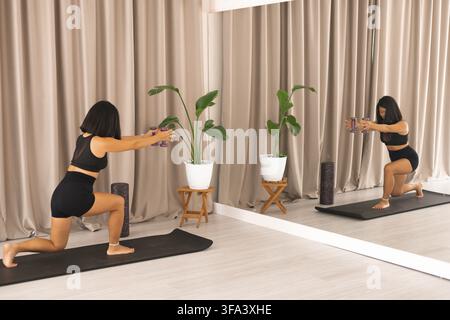 Una donna che fa fendenti con i manubri, specchiata davanti a lei, su un tappetino da yoga Foto Stock
