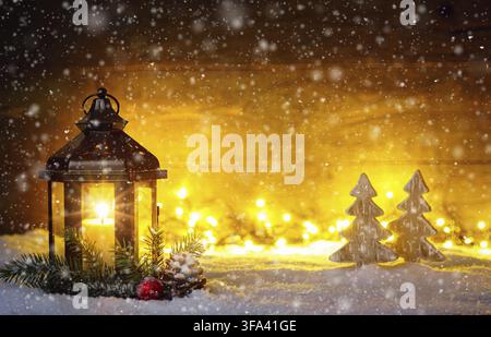 Scena natalizia con una lanterna, alberi, ramo di abete, fiocchi di neve e luci sfocate davanti a una tavola di legno scuro illuminata come spazio per il testo Foto Stock