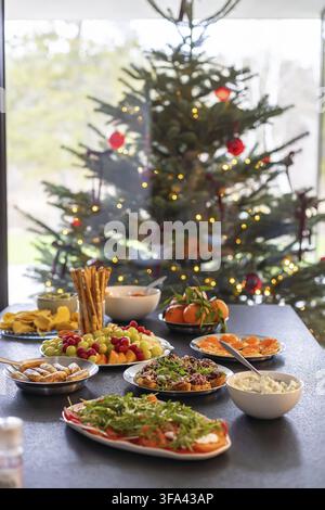 Un tavolo nero completamente apparecchiato con vari antipasti, frutta fresca, cracker e salse, posizionato davanti a un albero di Natale decorato vicino a una grande finestra Foto Stock