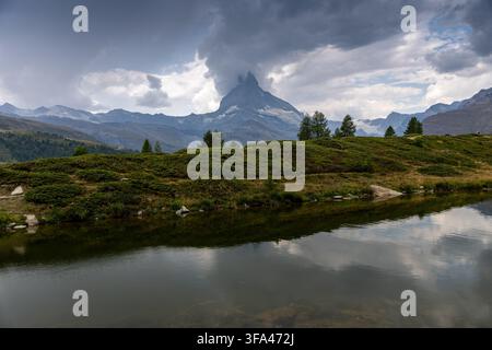 Il Matterhorn (Cervino) si erge sopra Zermatt nel Vallese, Svizzera, sotto un cielo estivo tempestoso. Il picco iconico si riflette in un tranquillo lago alpino Foto Stock