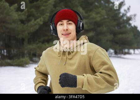Giovane uomo felice che fa jogging in una foresta innevata d'inverno con cuffie wireless e un berretto rosso. Ritratto di un corridore maschio che ascolta musica all'aperto durante Foto Stock