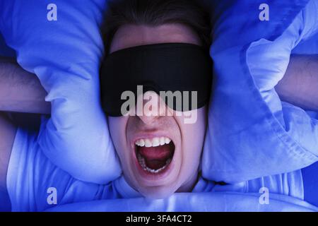 Un uomo arrabbiato che indossa una maschera per dormire urla nel letto mentre si copre le orecchie con i cuscini. L'immagine rappresenta l'insonnia, la mancanza di sonno o un nitrito rumoroso Foto Stock