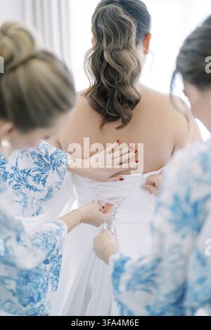 Primo piano di una sposa a cui due damigelle in abiti blu e bianchi allacciano il vestito da sposa bianco, messa a fuoco su mani e schiena Foto Stock