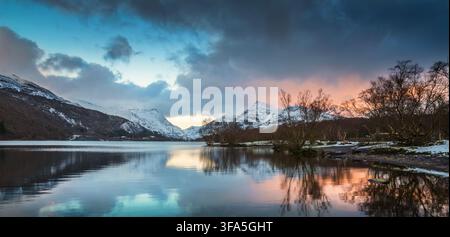 Un ampio tramonto invernale panoramico illumina nuvole pesanti sopra montagne coperte di neve che si riflettono nelle acque calme del Llyn Padarn nel Galles del Nord. Foto Stock