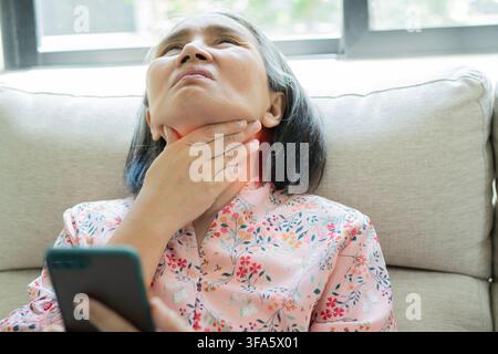 Donna asiatica matura seduta sul divano con dolori al collo mentre usa uno smartphone a casa. Concetto di assistenza sanitaria, stile di vita e tecnologia Foto Stock