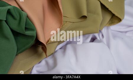 Vista ravvicinata intima di tessuti premium a strati che mostrano tessuti verde vivace, delicato pesca e caldo kaki che riposano dolcemente su un letto di immacolato Foto Stock
