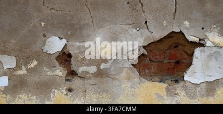 Vista ravvicinata orizzontale di una superficie murale in decadenza con strati di vernice gialla e bianca su cemento crepato e mattoni rossi usurati e malta esposti Foto Stock