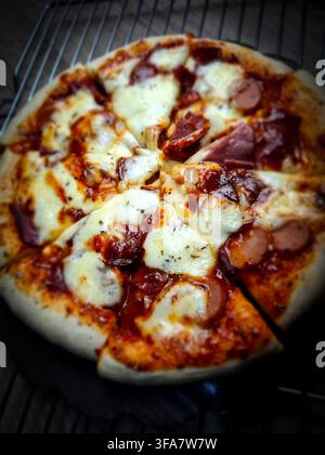 Primo piano di una pizza fatta in casa appena sfornata con formaggio mozzarella fuso, fette di salsiccia e sugo di pomodoro saporito. La pizza è posata su una griglia di raffreddamento, hig Foto Stock