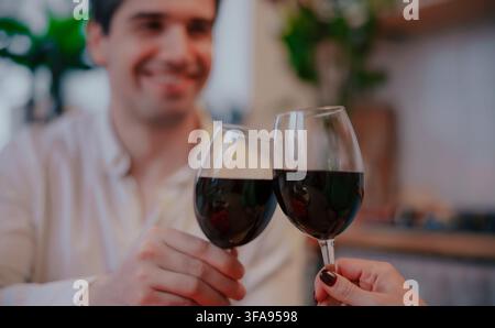 Due bicchieri di vino rosso si incontrano in un brindisi celebrativo mentre un uomo sorridente rimane leggermente sfocato sullo sfondo in un ambiente domestico accogliente. Foto Stock